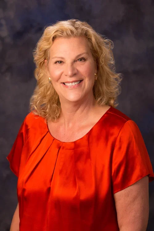 Woman with blond curly hair wearing a shiny red blouse smiles at the camera