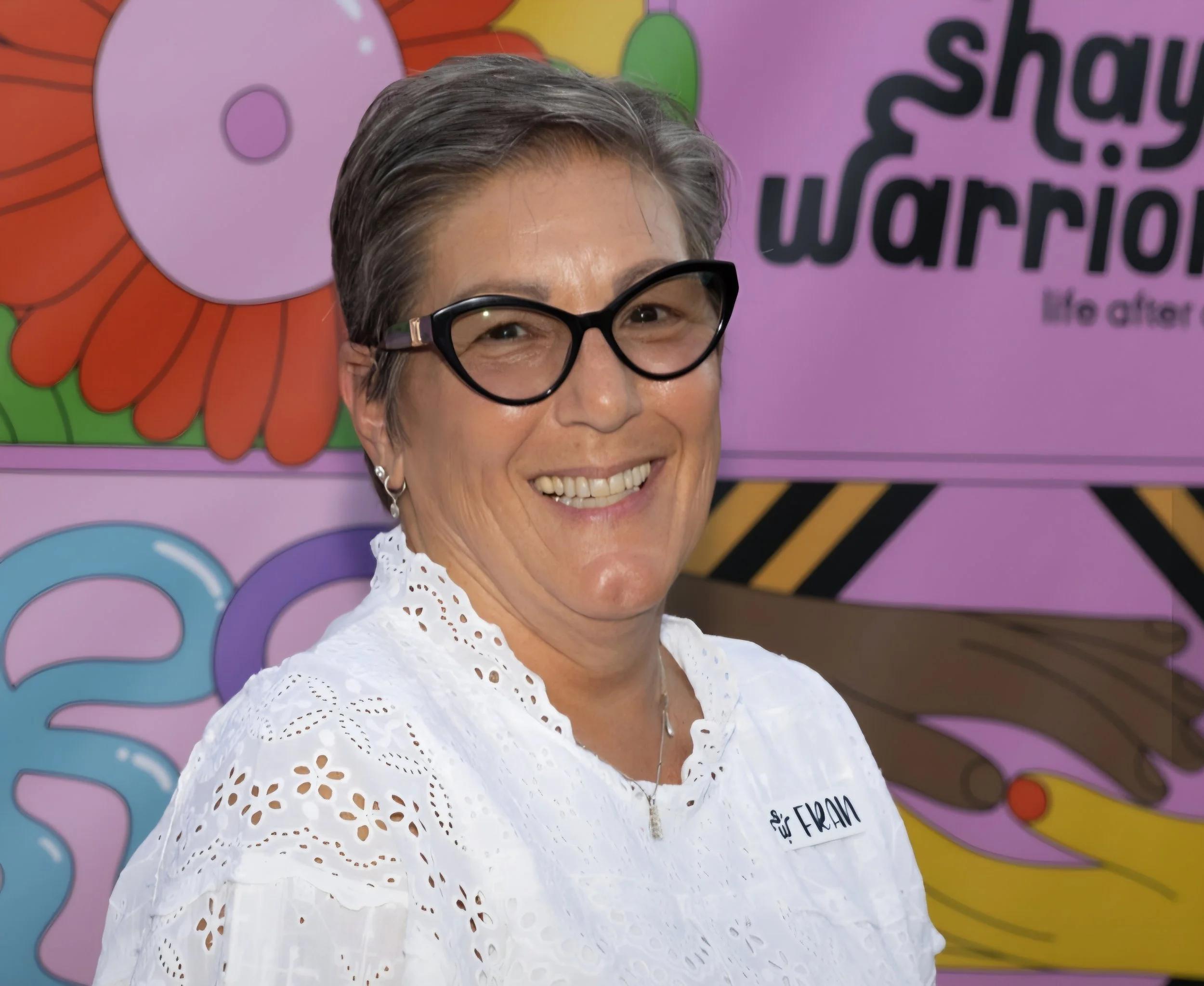 A smiling woman wearing black glasses and a white eyelet blouse, standing in front of colorful background with large flower and question mark illustrations and text.