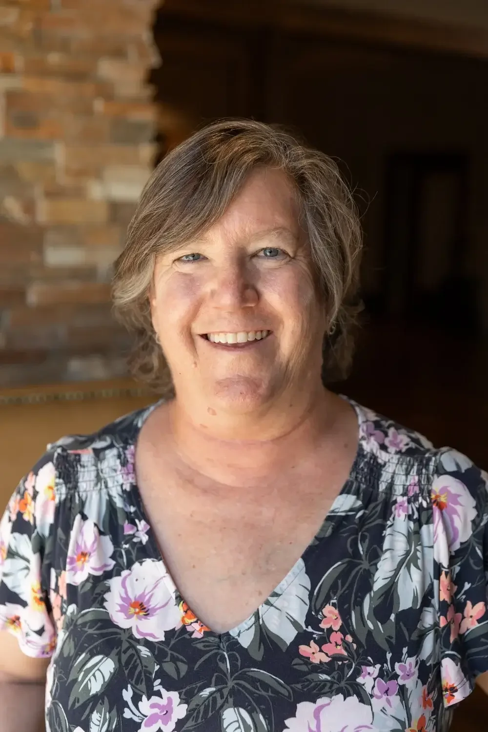 A smiling woman wearing floral patterned blouse, standing in front of a brick wall.