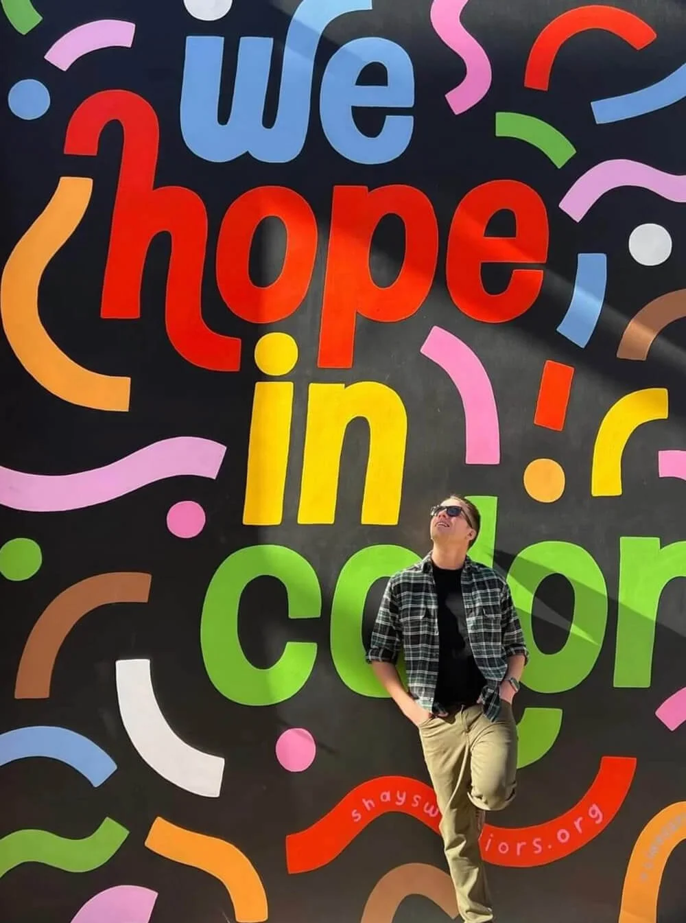 A man stands with one foot against a large mural looking up at the words. The words on the mural read we hope in color.