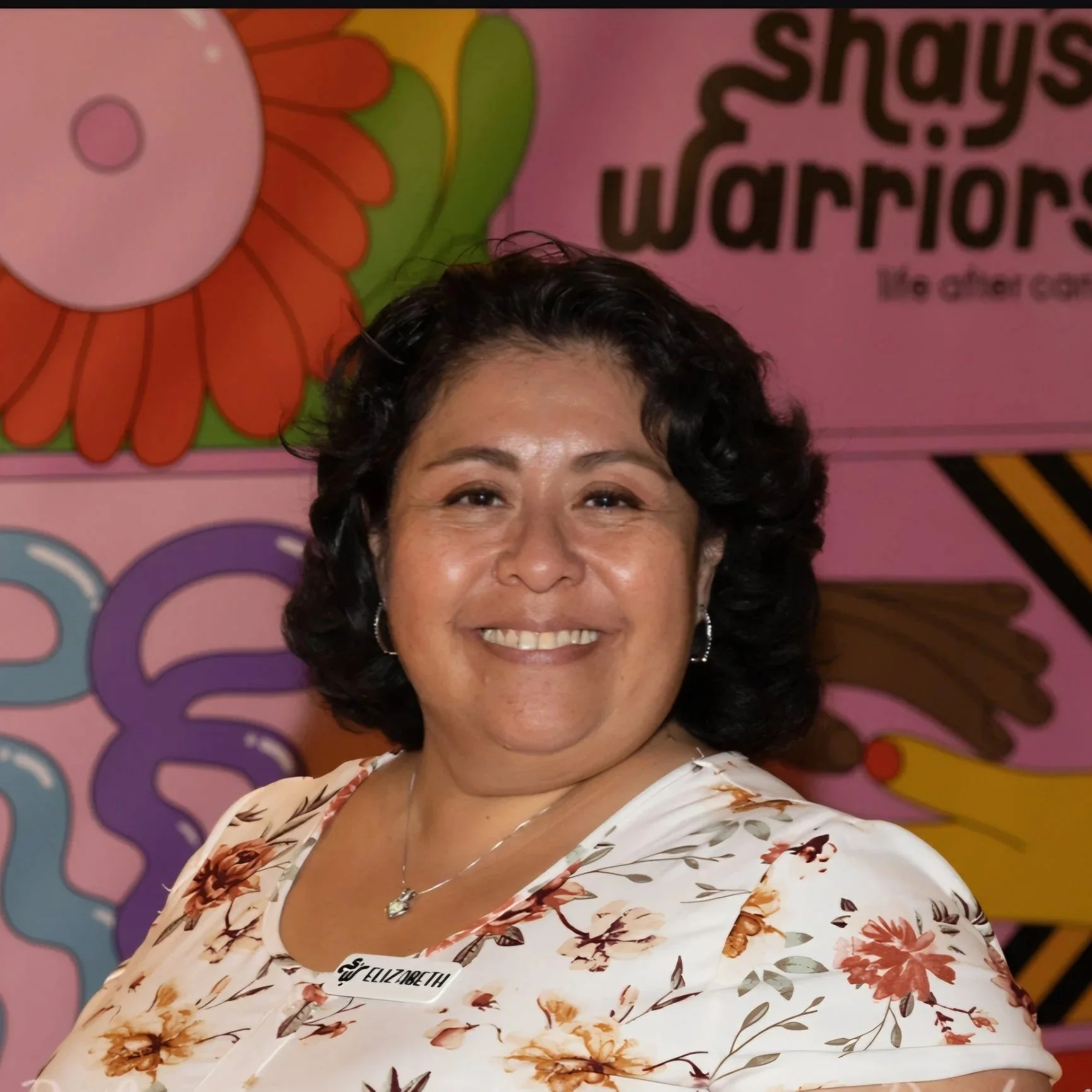 Smiling woman with short black hair wearing a floral dress, necklace, earrings, and a name tag that says Elizabeth, in front of colorful background with a large pink flower and the words 'Shays Warris'