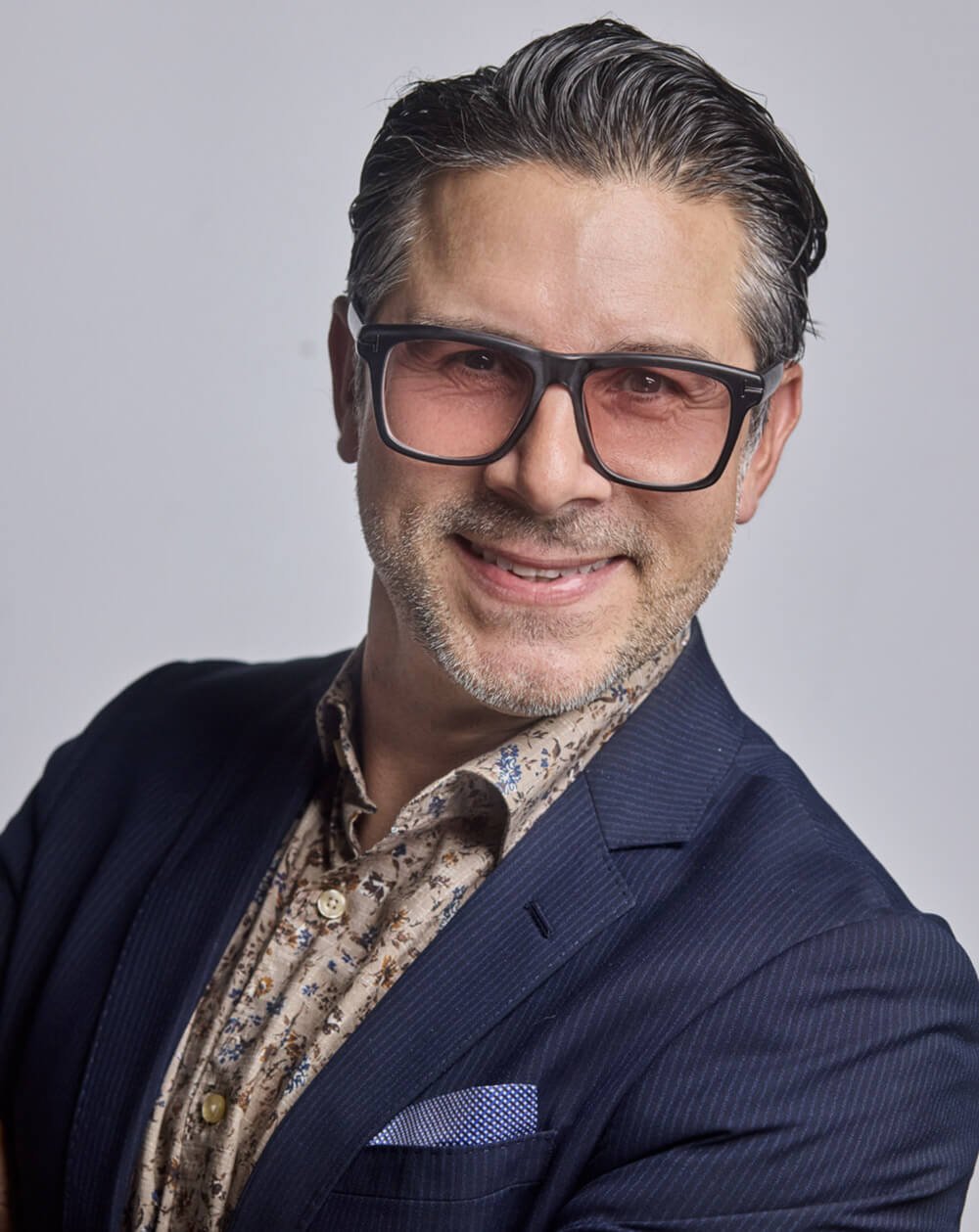 A middle-aged man with dark, slicked-back hair, wearing large black glasses, a navy suit jacket, a patterned button-up shirt, and a pocket square, smiling at the camera against a plain gray background.