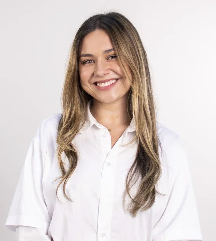 Woman with long light brown hair wearing a white long sleeved button up shirt smiles at the camera