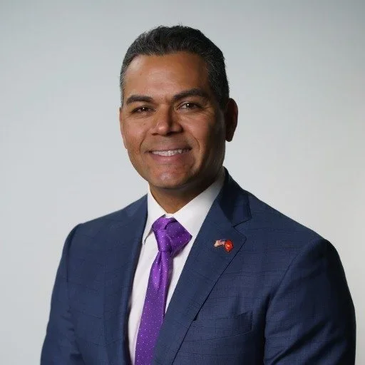 Man with short dark hair wearing a blue suit jacket, white dress shirt, and a purple tie