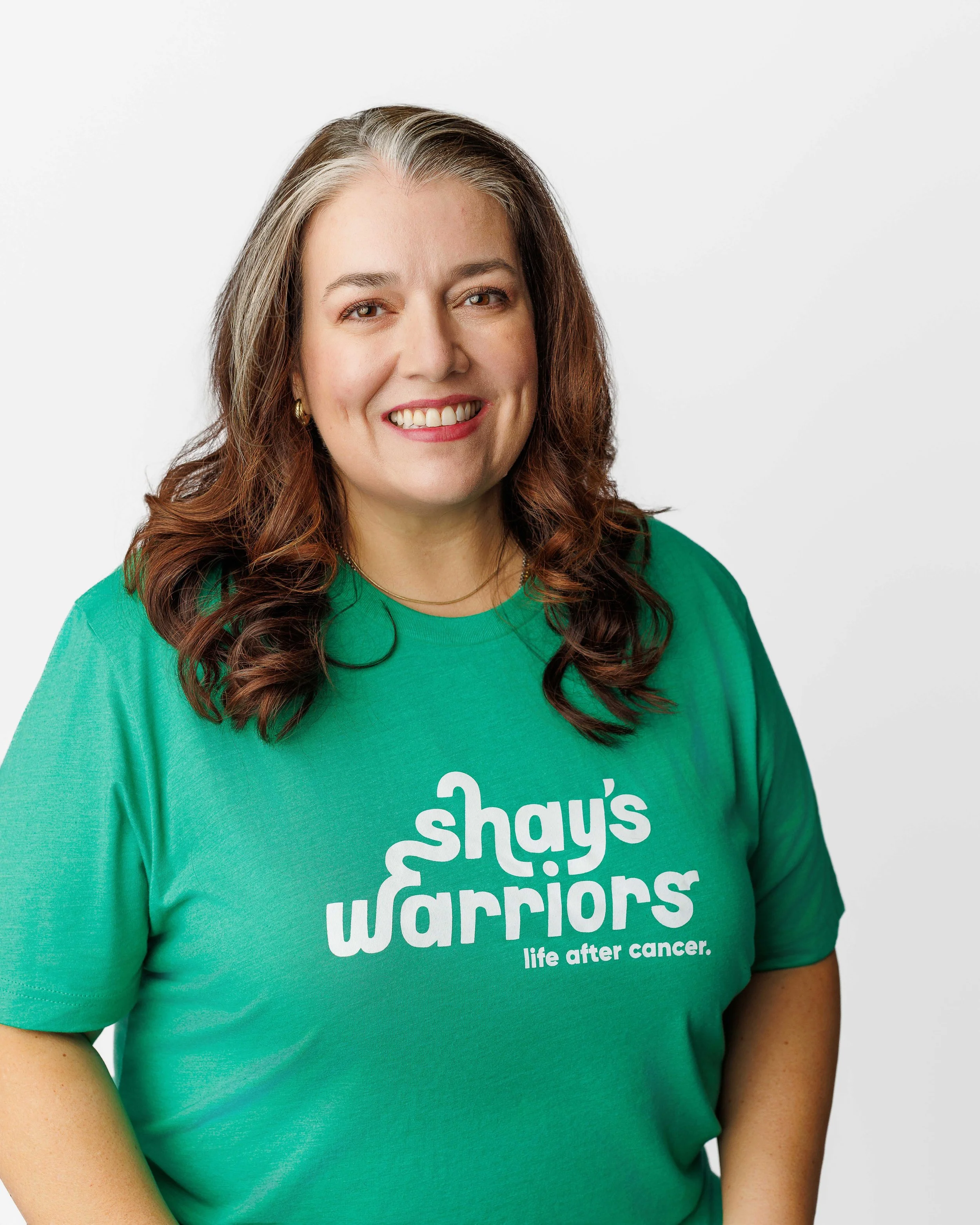 Close-up of a smiling woman wearing a green shirt with white text supporting Shays Warriors for life after cancer.
