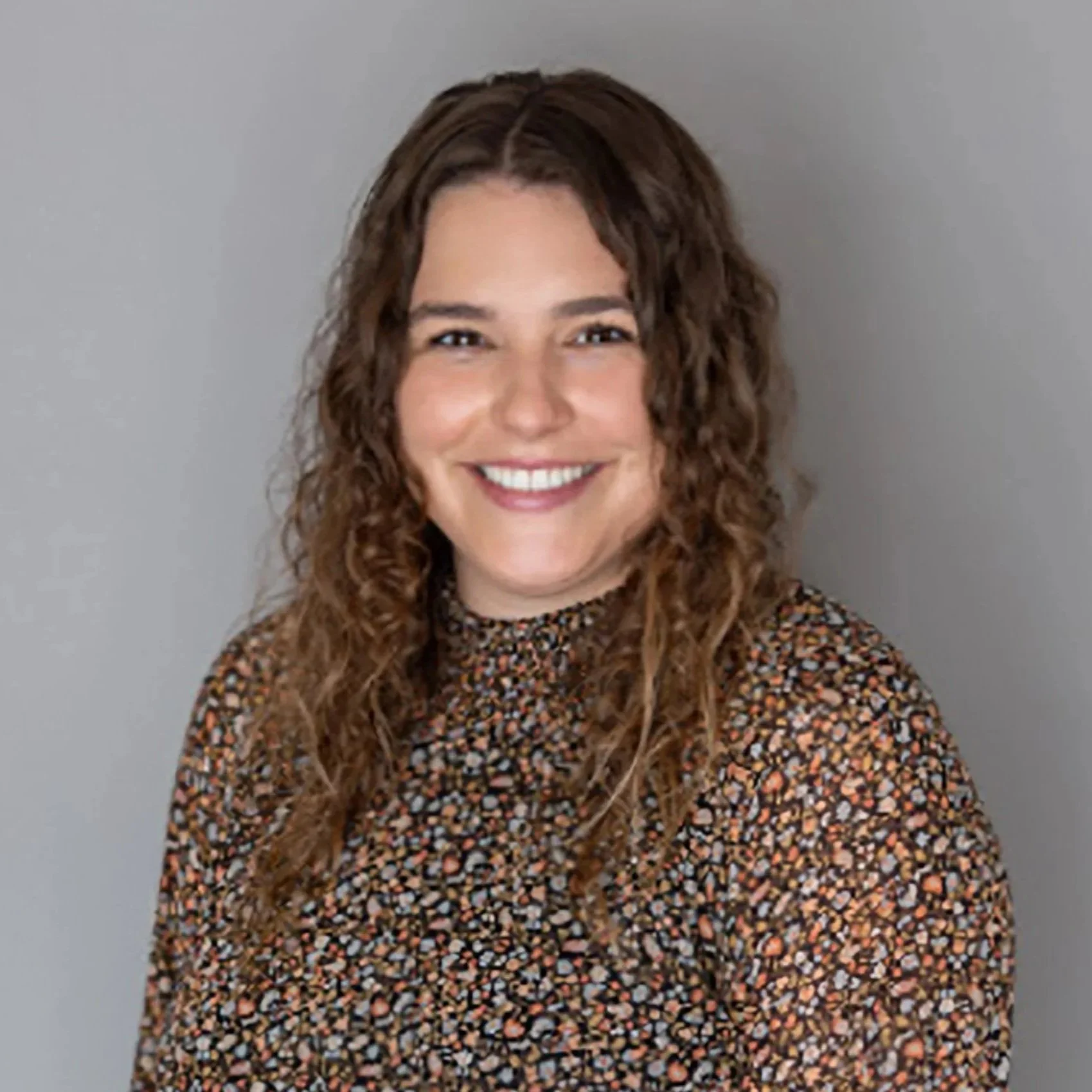 Portrait of a woman with curly brown hair, smiling, wearing a floral patterned top, against a plain gray background.