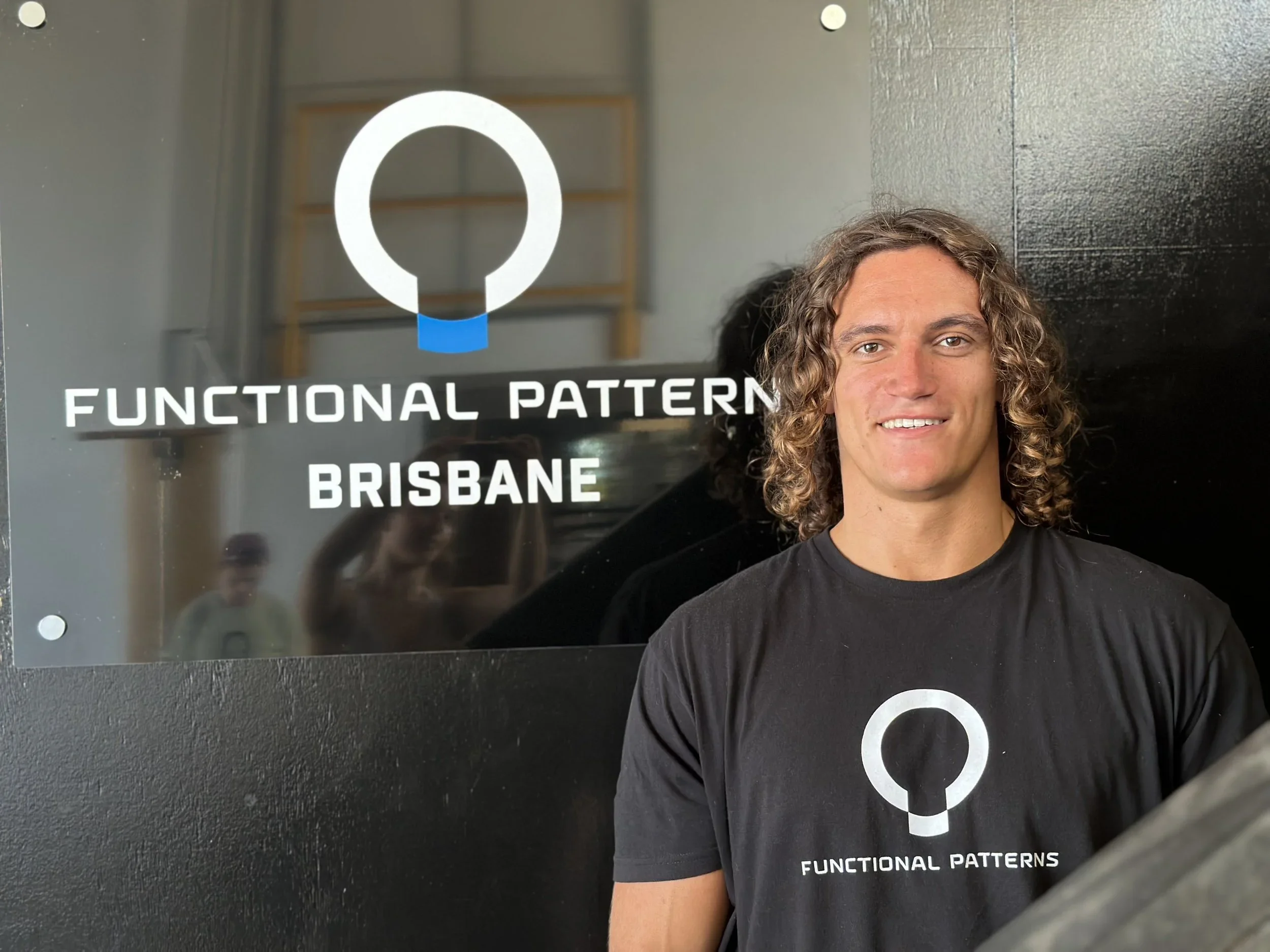 A young man with curly hair wearing a black T-shirt standing in front of a sign that reads 'Functional Patterns Brisbane' with a logo above and to the left of the text.