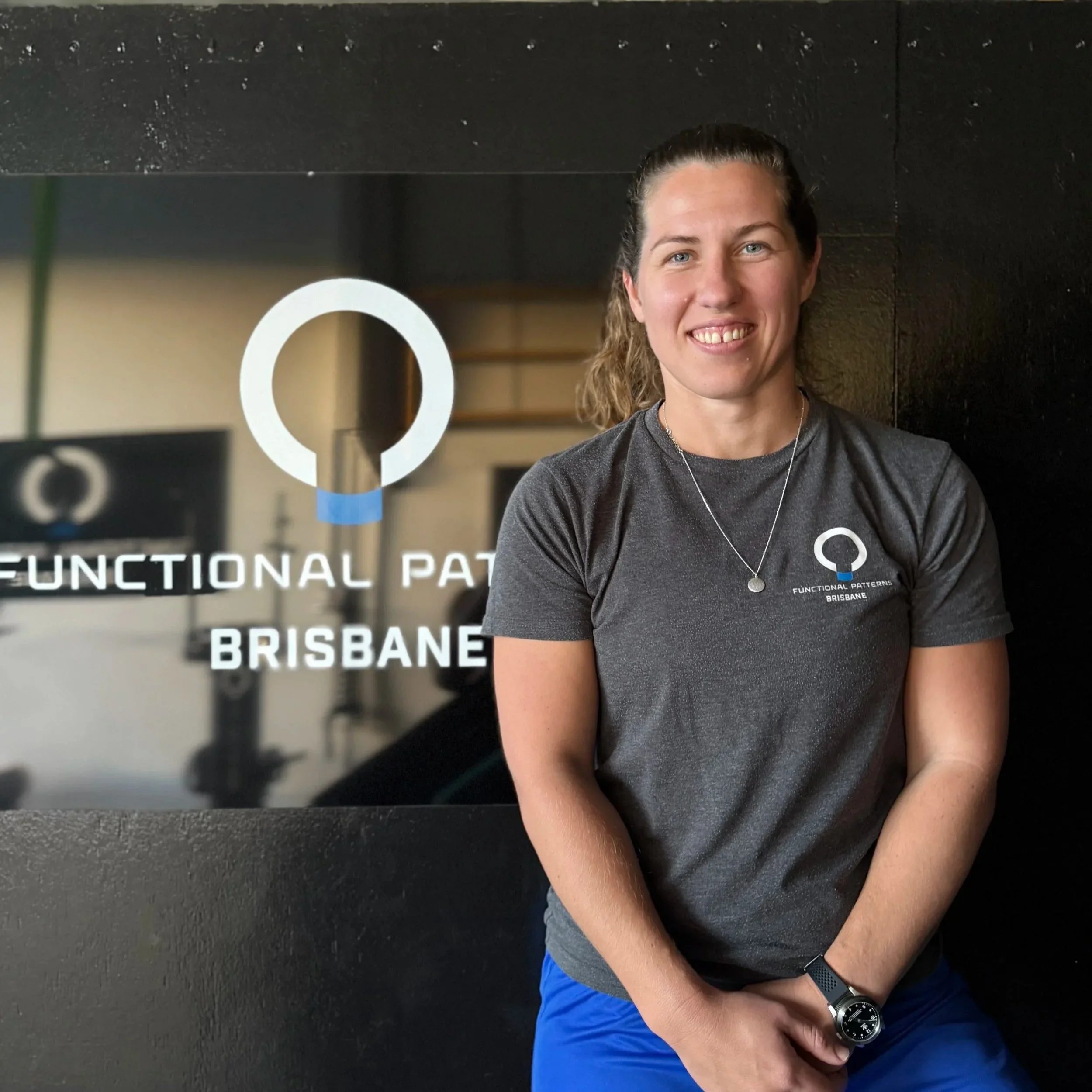 A woman in a gray T-shirt and blue shorts standing in front of a logo and text that reads "Functional Patterns Brisbane". She is smiling, with her hair tied back, and wearing a watch and a necklace.