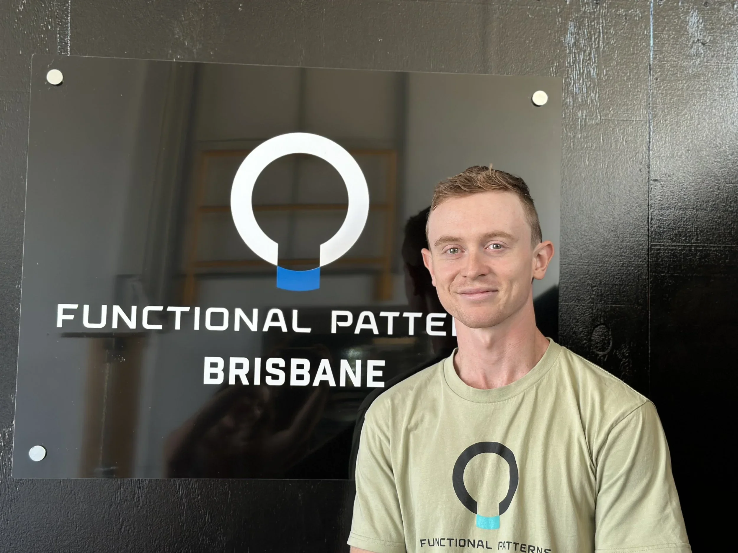 A man with short light brown hair and a light green t-shirt standing in front of a sign that reads 'Functional Patterns Brisbane' with a logo above the text.