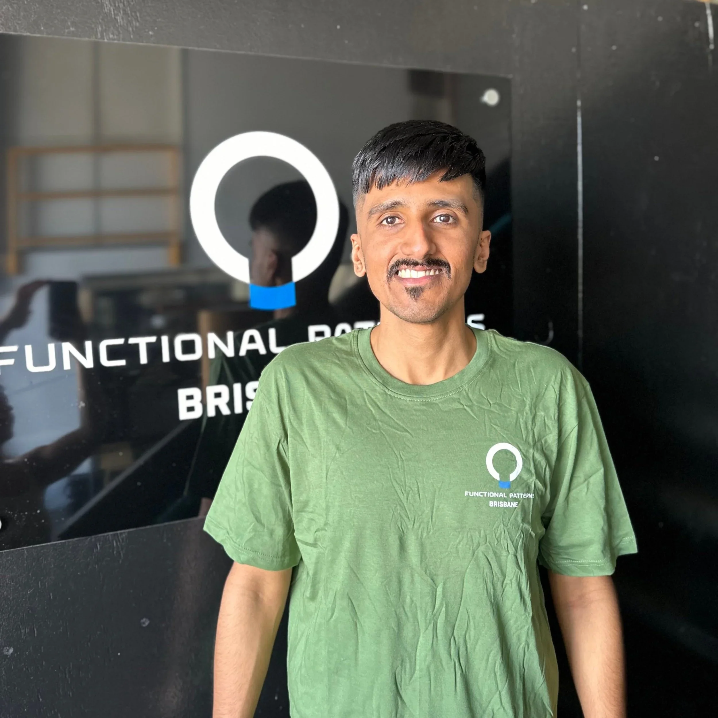 A young man with a short beard and mustache, wearing a green T-shirt with a logo and text, smiling and standing in front of a black wall with a logo and white text.
