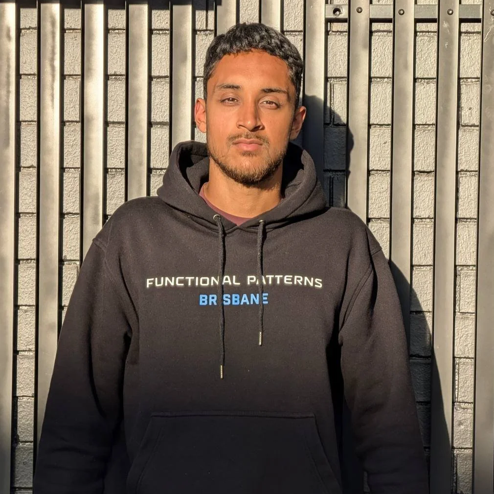 A man with short dark hair and a beard standing against a brick and metal fence, wearing a black hoodie with white and blue text that reads "Functional Patterns Brisbane."