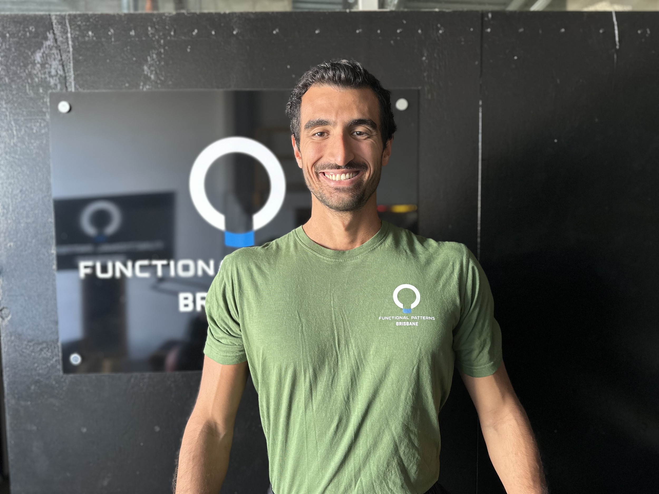 A smiling man with dark hair and a beard wearing a green t-shirt standing in front of a glass sign that reads 'Functional Patterns Brisbane' with a logo above it.