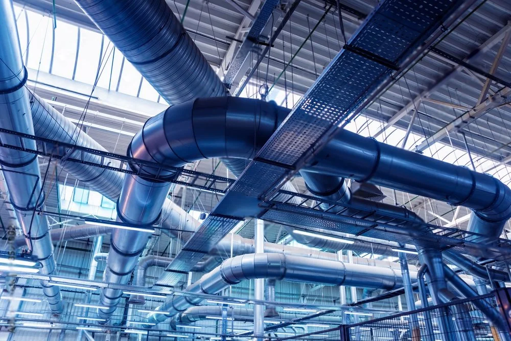 Industrial ductwork and piping system inside a large factory or plant with metal framework and skylights.
