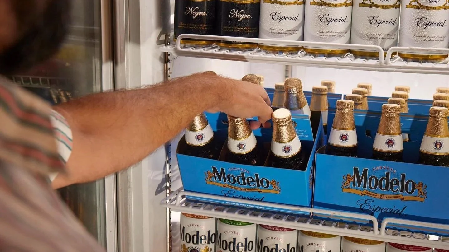 a man reaches into a cold display to grab a 6-pack of Modelo in glass bottles