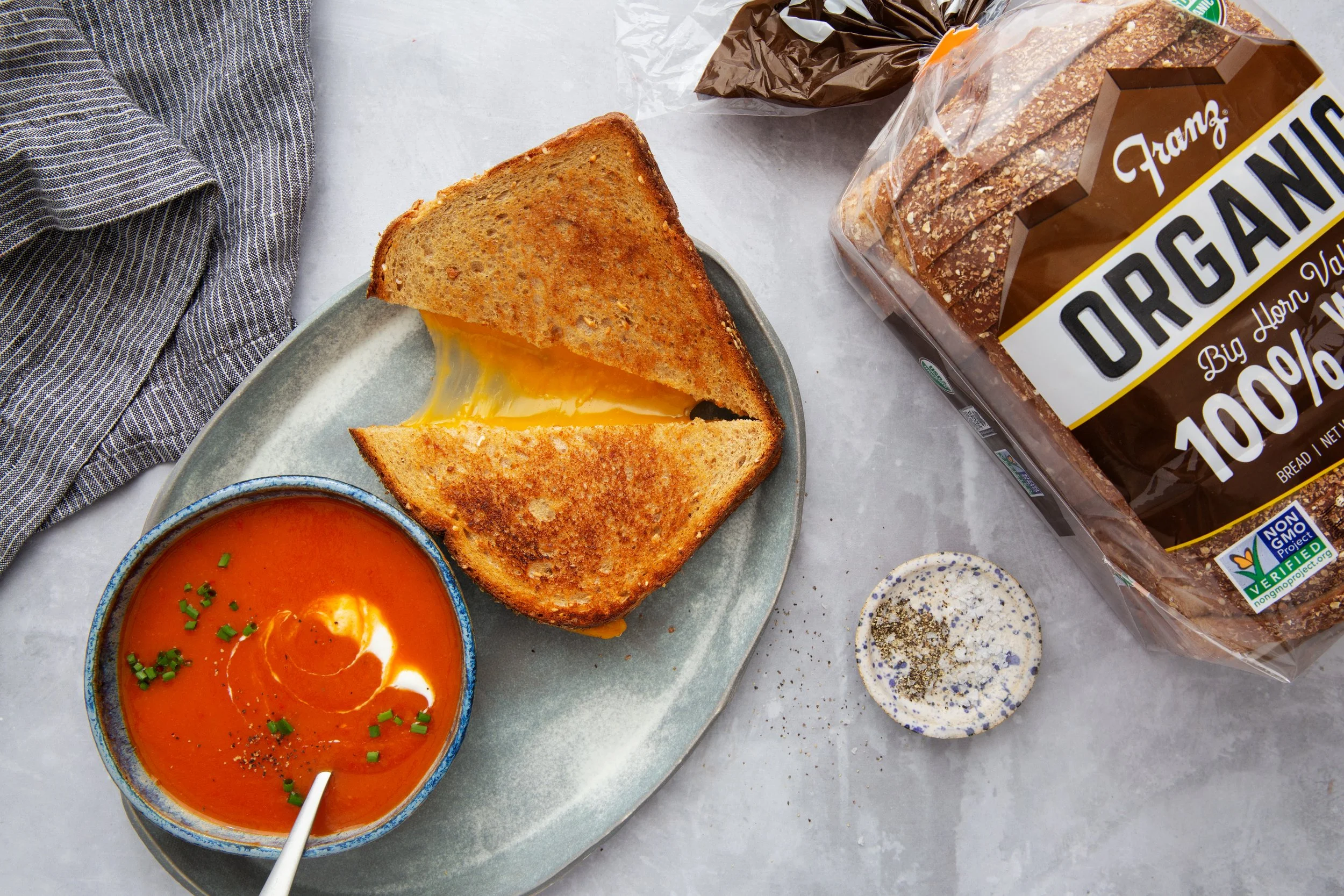 Grilled cheese sandwich with cheddar cheese pull, next to a bowl of tomato coup, on a stone oval dish, next to a package of Franz Organic 100% Wheat Sliced Bread