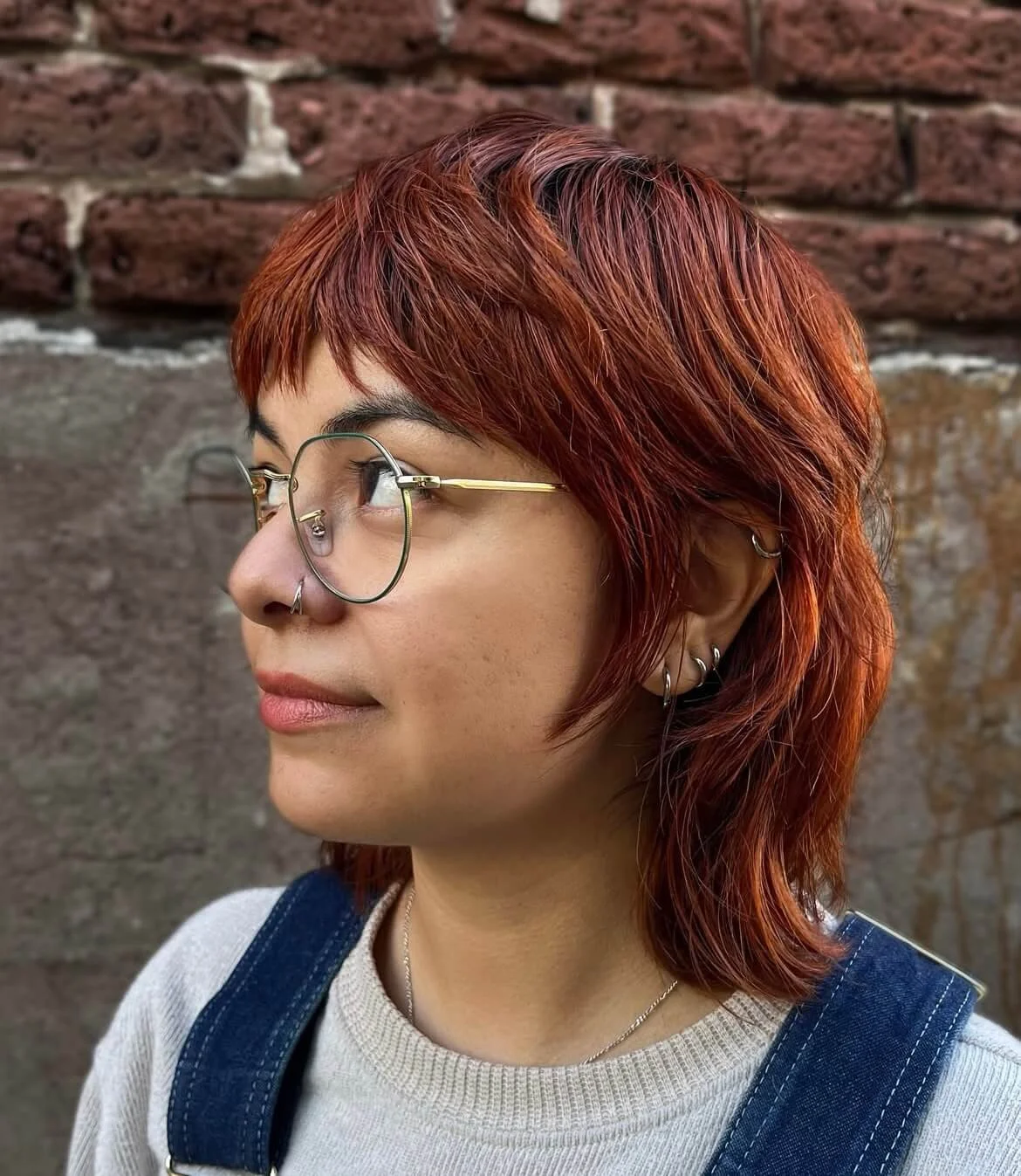 Side profile of a woman with short red hair, wearing glasses and earrings, standing against a brick wall.
