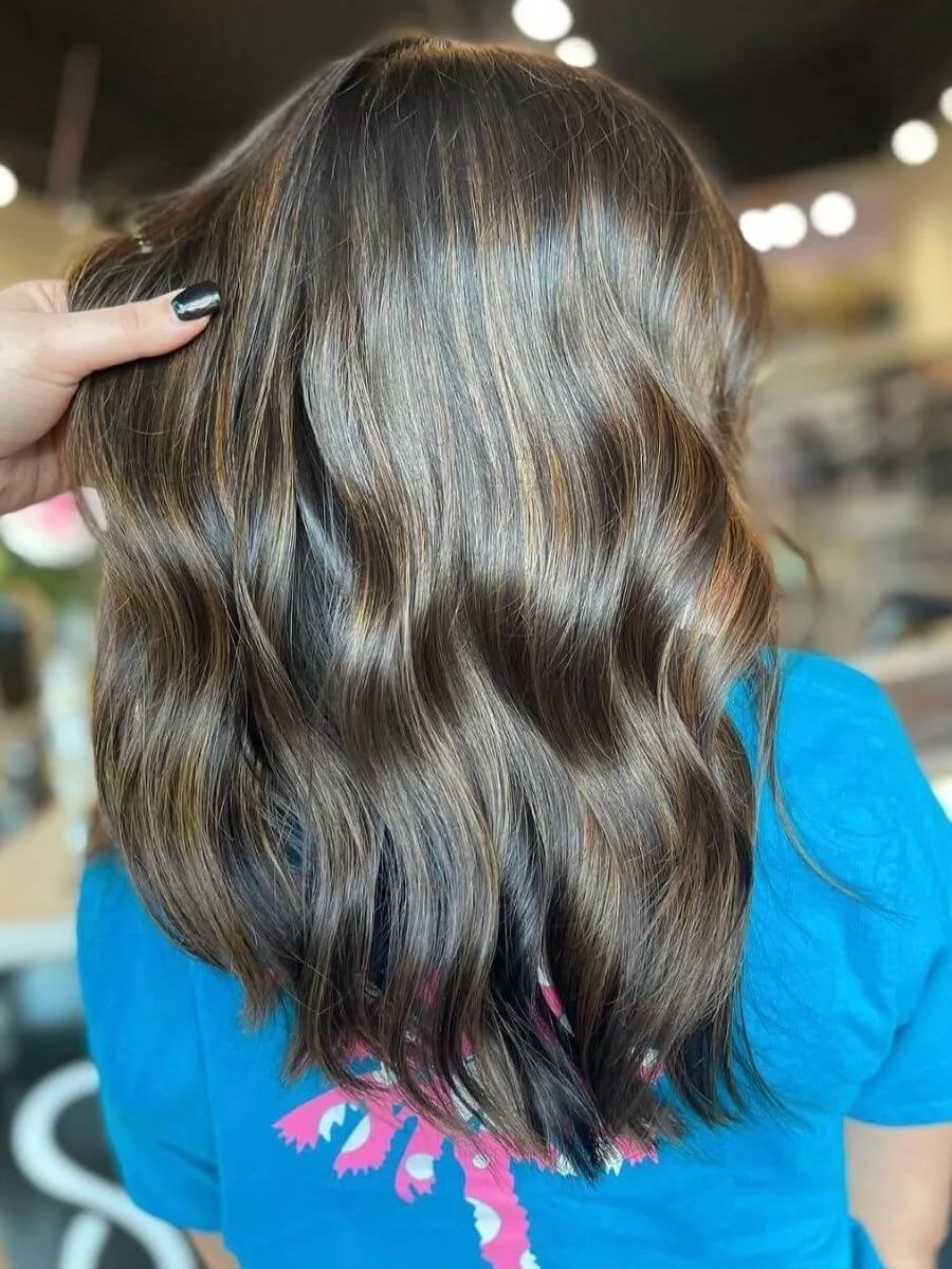 Person with shoulder-length wavy brown hair, viewed from behind, wearing a blue shirt with pink and white design.