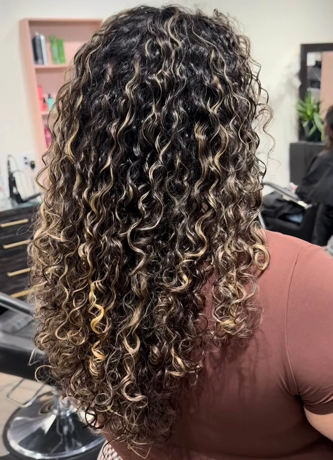 Close-up of a woman’s head with long, curly, highlighted hair, sitting in a salon. The background includes salon furniture and a stylist.