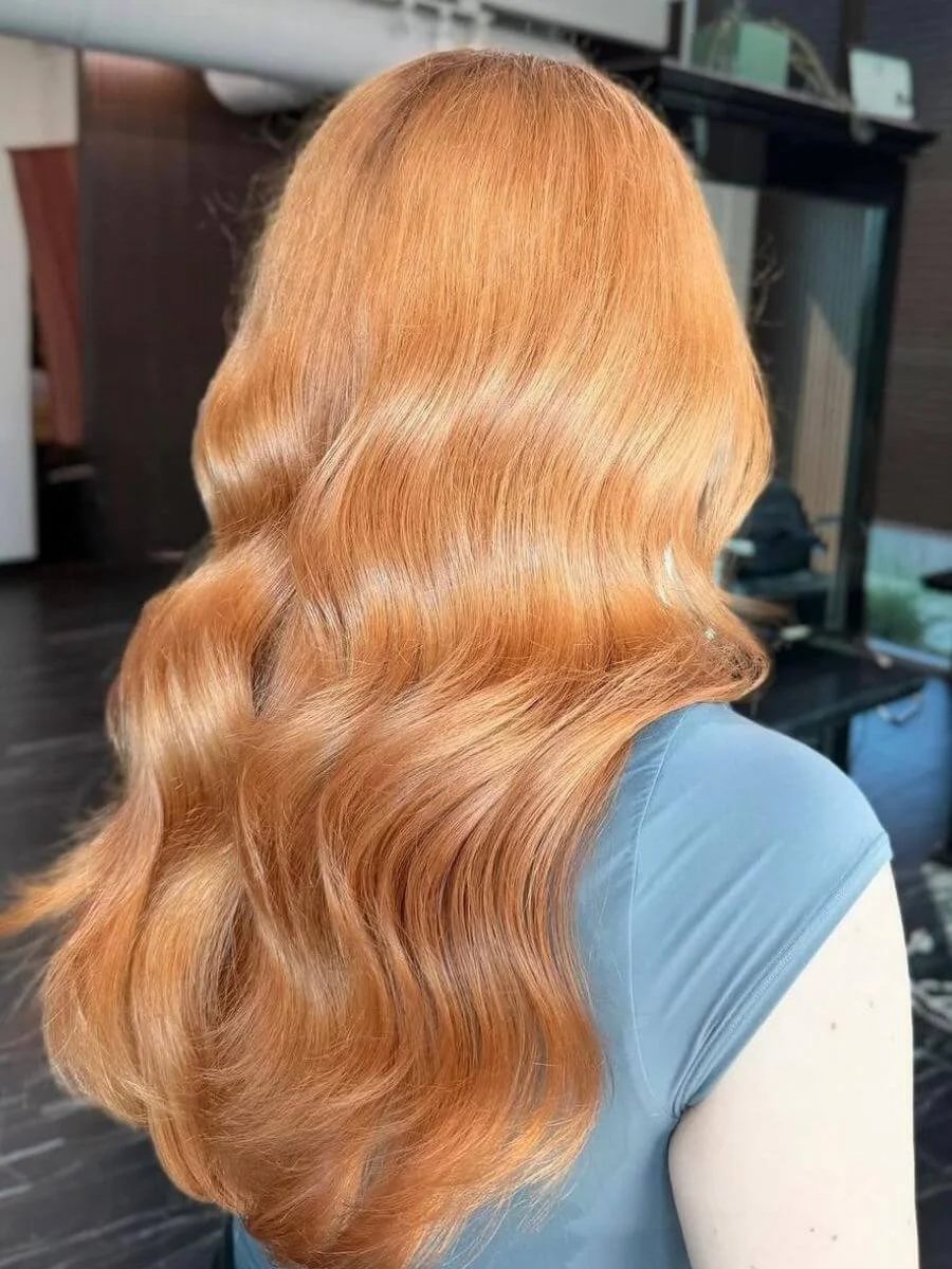 Side view of a woman with long, wavy, strawberry blonde hair in a salon or indoor setting.