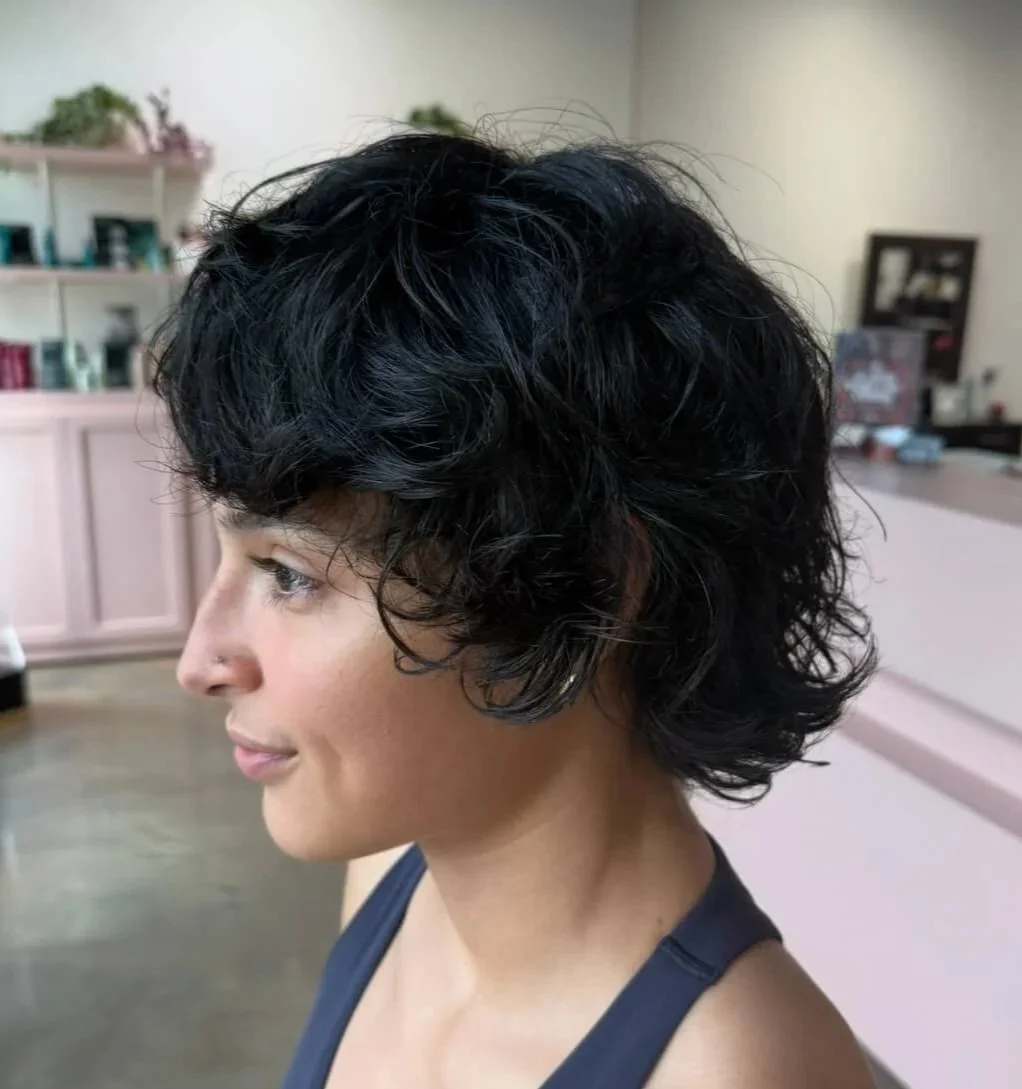 Side profile of a woman with short, curly black hair in a room with pastel pink cabinets and shelves in the background.