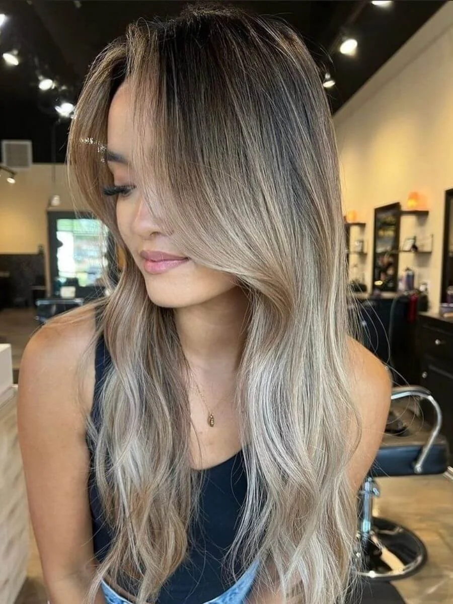 A woman with long, balayage blonde hair styled in loose waves, posing in a salon with black chairs and shelves in the background.