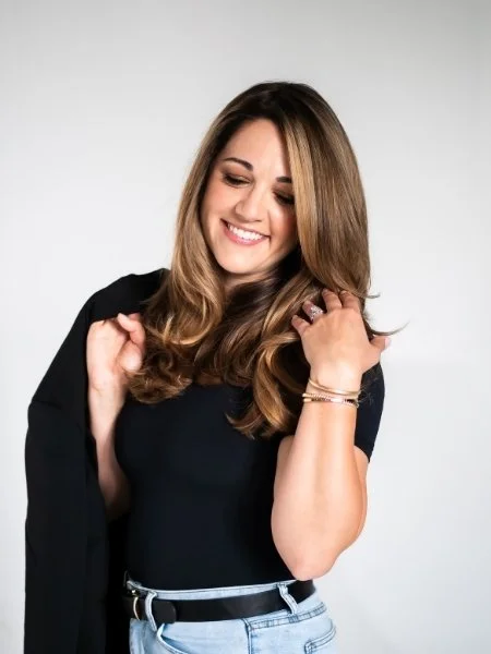 A young woman with long brown hair smiling with closed eyes, wearing a black top, light blue jeans, and a black jacket draped over her shoulder, posing against a plain light gray background.