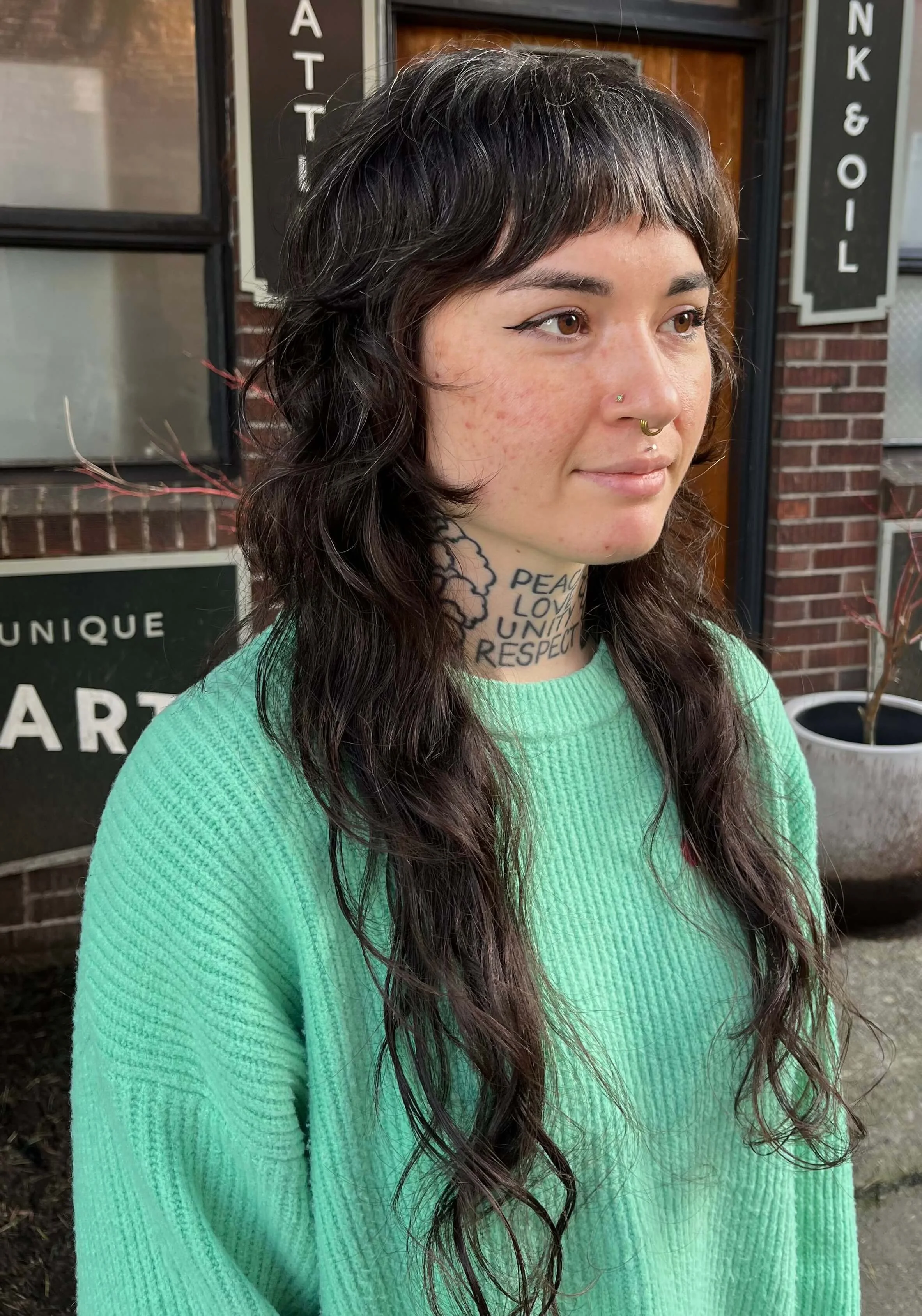 A woman with dark wavy hair and multiple facial piercings, wearing a light green sweater, standing outside in front of a brick building with various signs.