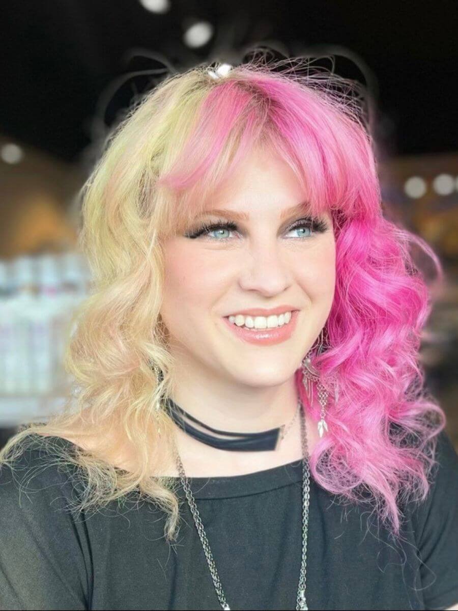 A smiling woman with blonde and pink curly hair, wearing a black top, a choker, and earrings, against a blurred indoor background.