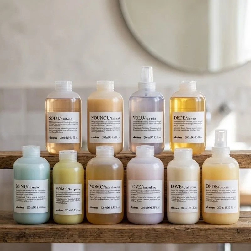Collection of ten bottles of hair care products on a wooden shelf in a bathroom, including shampoo, conditioner, hair mist, and hair masks in various pastel and neutral colors.