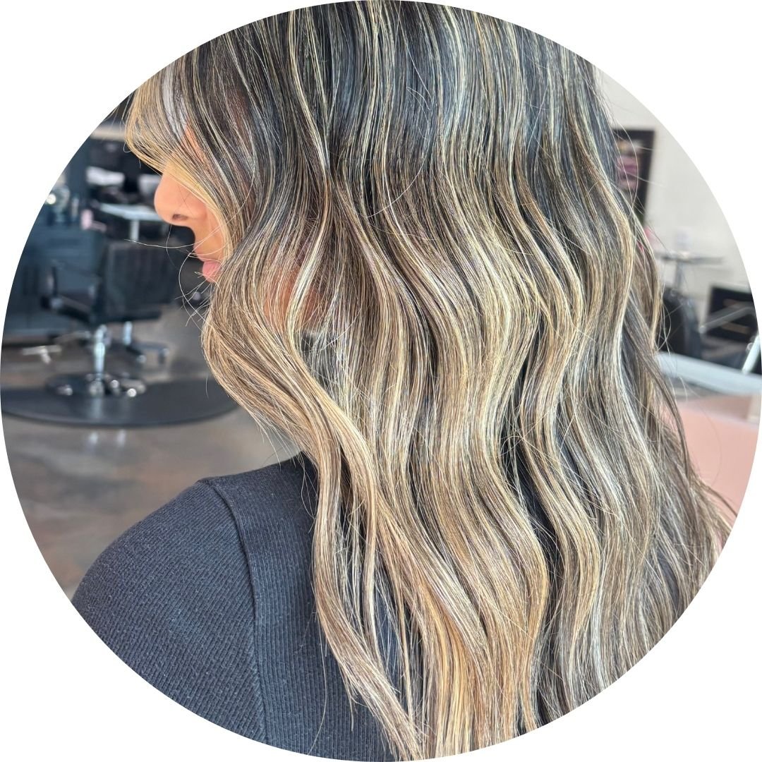 A woman with long, wavy hair styled in blonde highlights and lowlights, wearing a black top, at Spoleti Salon in Austin.