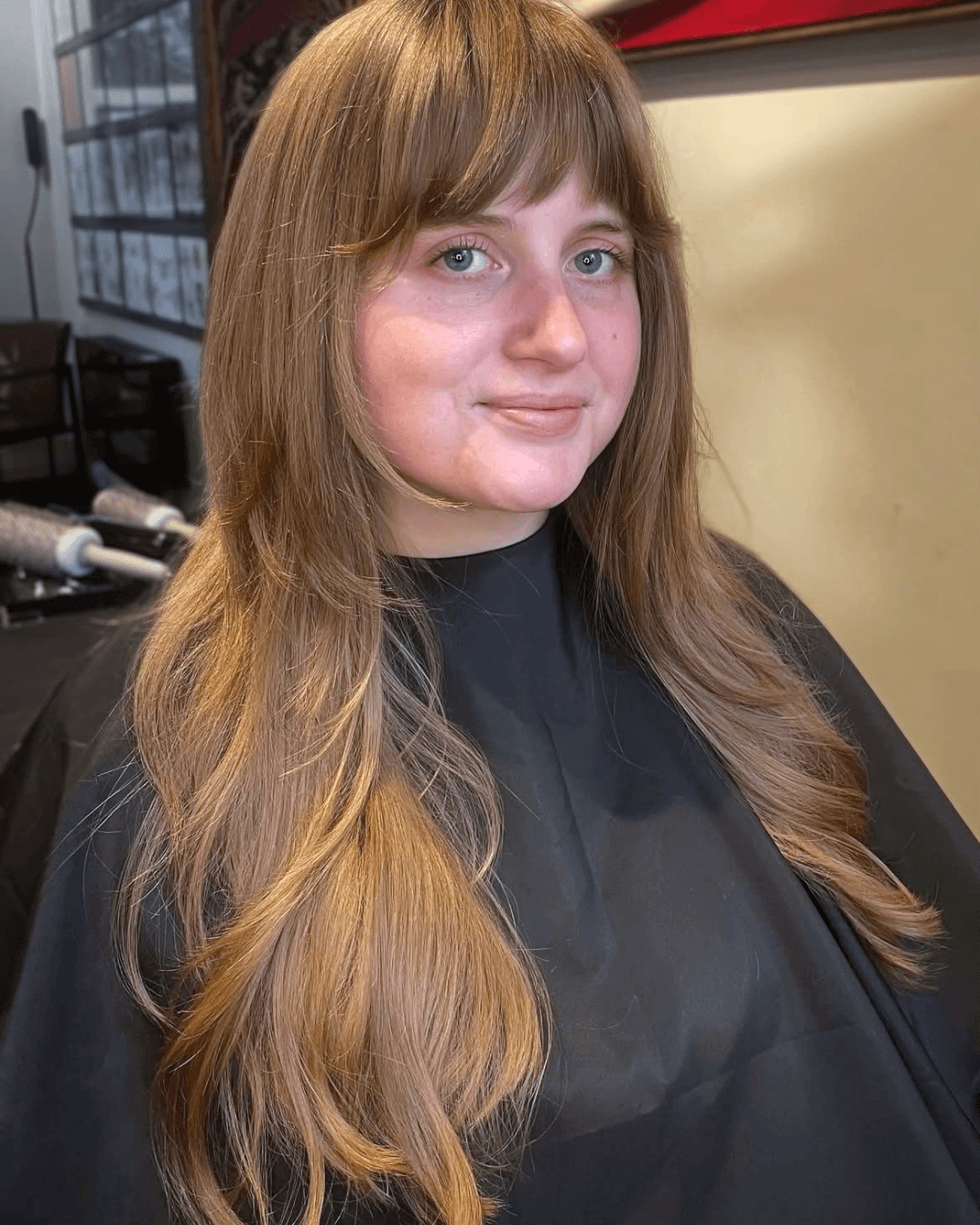A woman with long, wavy auburn hair and blue eyes sitting in a salon chair, wearing a black cape, with salon equipment in the background.
