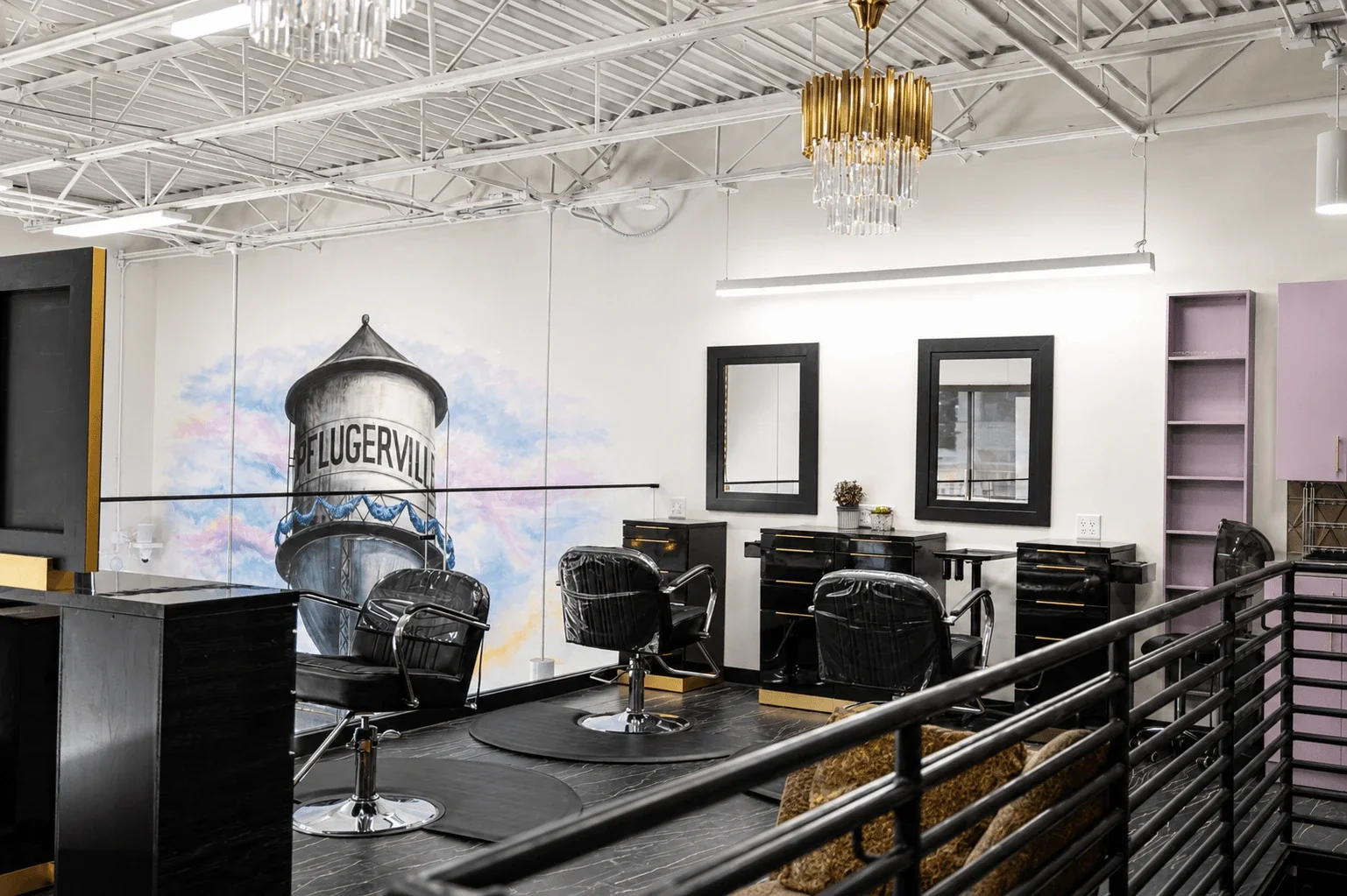 Modern salon or barbershop interior with black chairs, black furniture, a wall mural of a water tower with the text "OF LUGERVILLE," and decorative mirrors with black frames, illuminated by a gold chandelier and linear lighting.