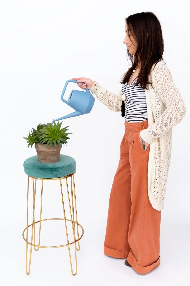 Woman watering a potted plant placed on a small green velvet stool with a gold metal frame, against a plain white background.