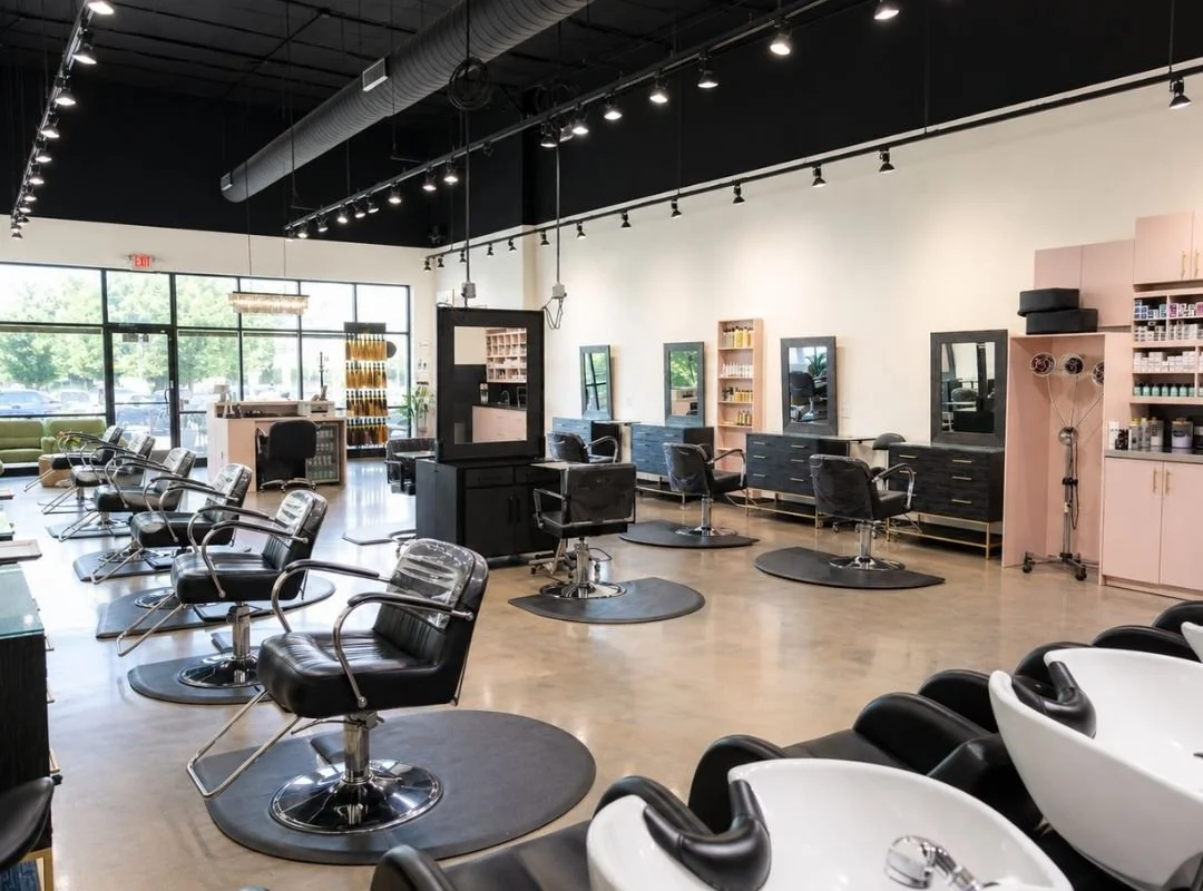 Empty modern hair salon with black and white styling chairs, mirrors, and hair products on shelves, illuminated by track lighting.