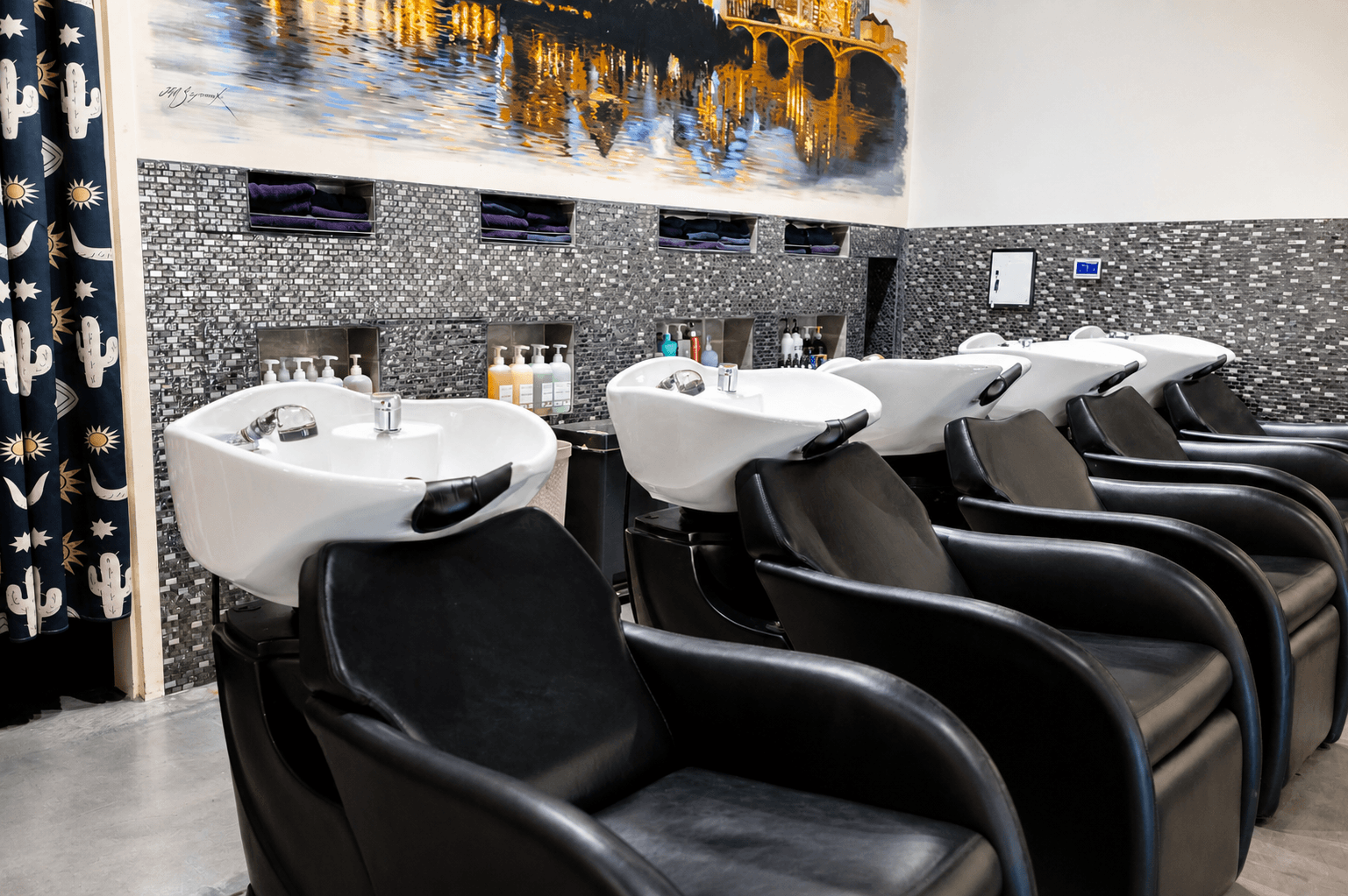 Salon with five black shampoo chairs and white washing stations, decor includes a mural of a cityscape under a bridge and western-themed curtains.