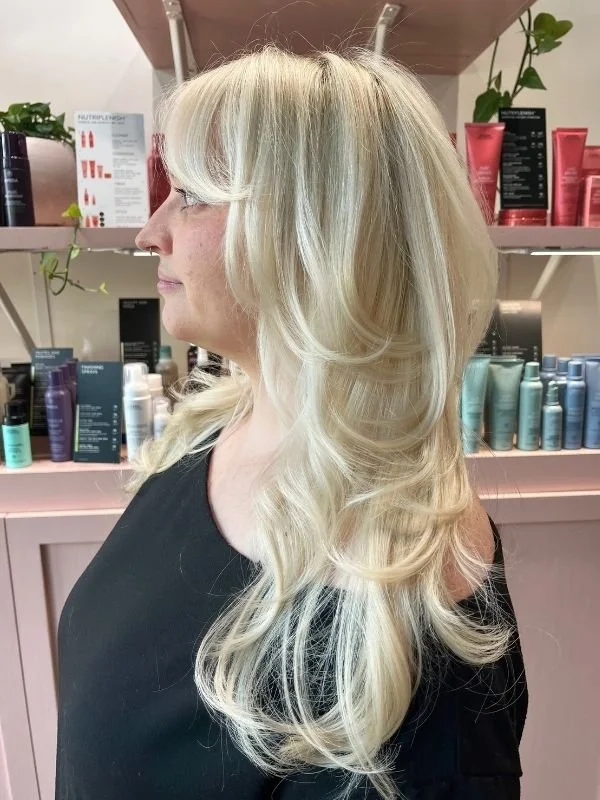 Profile view of a woman with long, platinum blonde, styled wavy hair in a salon setting with hair products on shelves behind her.