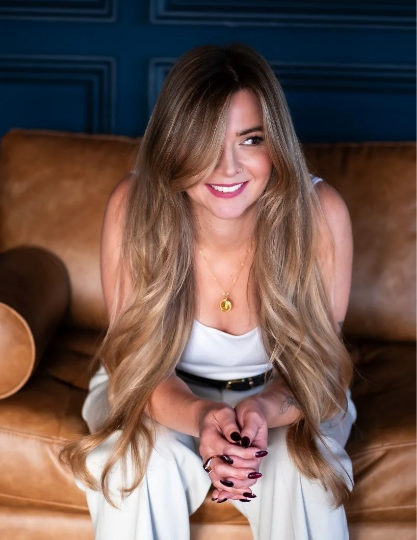 A smiling woman with long wavy hair, wearing a white outfit, gold necklace, and dark lipstick, sitting on a brown leather sofa with a blue wall in the background.