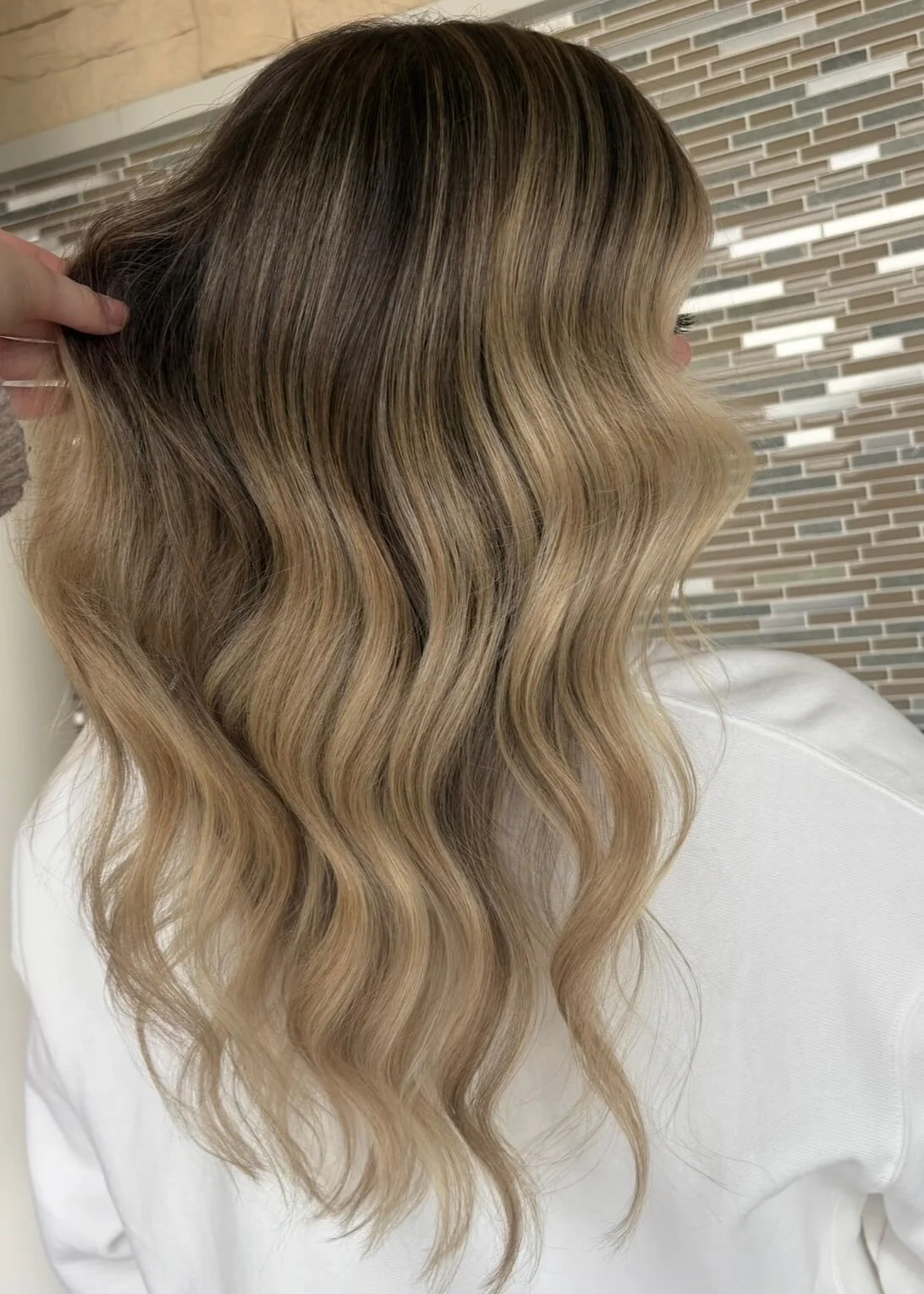 Woman with blonde, wavy hair facing sideways and wearing a white shirt, with a tiled wall in the background.