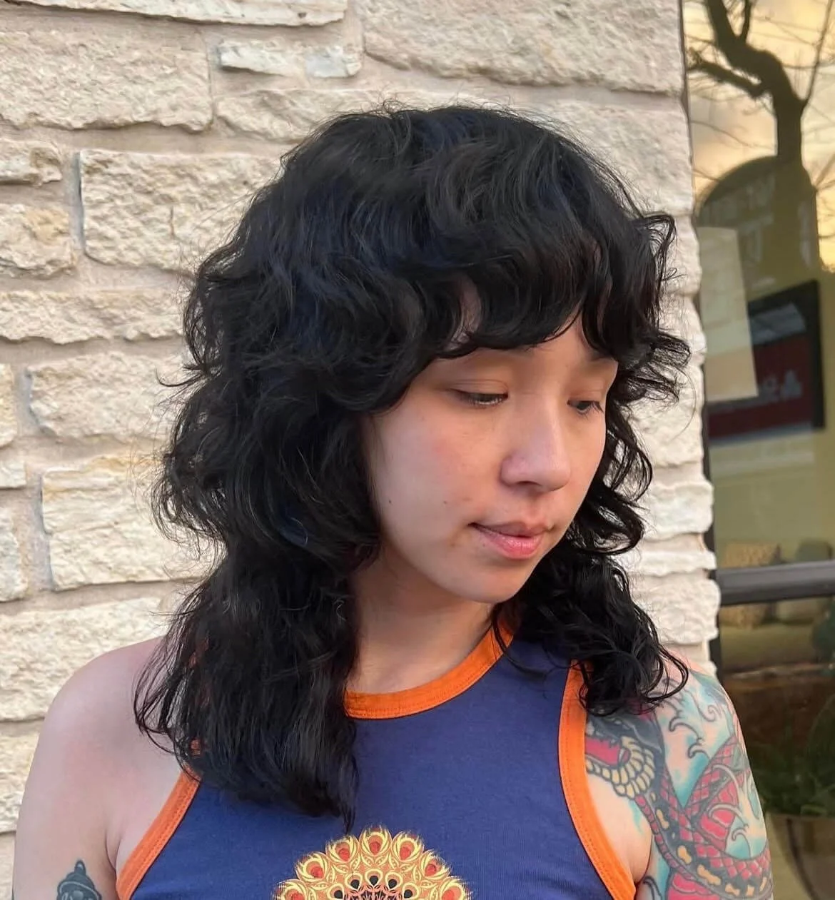 A young person with curly black hair, wearing a blue sleeveless shirt with orange trim, standing outdoors against a brick wall background, looking downward.