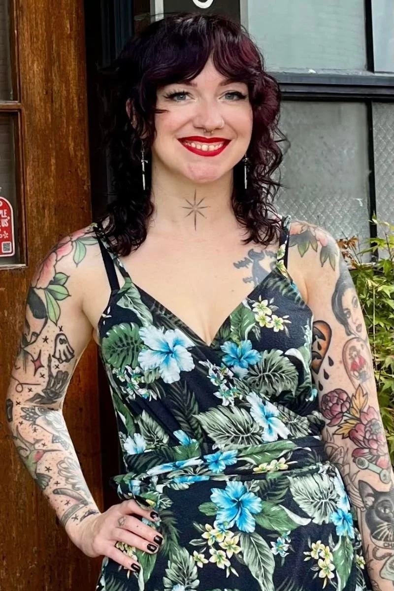 Woman with dark purple, curly hair wearing a black floral dress, standing outdoors near a wooden door and window, with visible tattoos on her arms and chest.