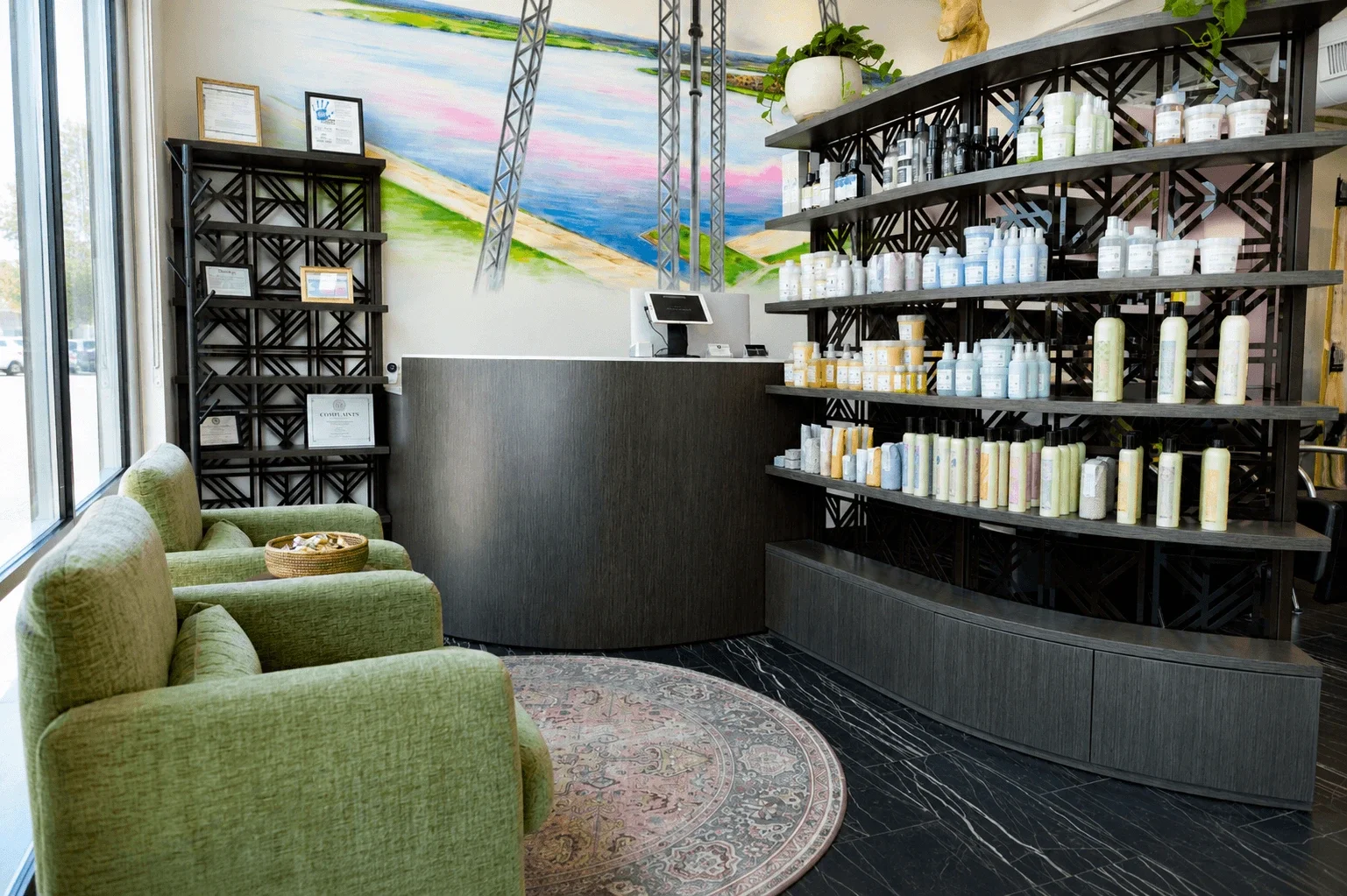Inside a spa or wellness center reception area with green armchairs, a dark marble floor, black shelves filled with bottled products, a small curved reception desk with a computer, large window on the left, and a colorful mural on the wall behind the counter.