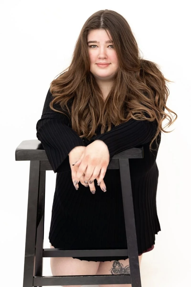 A young woman with long, wavy brown hair sitting on a black stool, wearing a black ribbed sweater, with her hands resting on the stool and a tattoo of a butterfly visible on her thigh.
