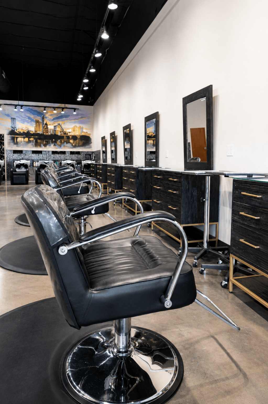 Row of black leather salon chairs with chrome arms in a modern hair salon, with mirrors and black cabinetry along the wall, and a large mural of a city skyline on the back wall.