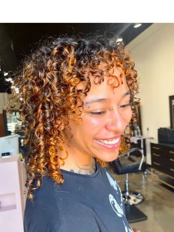 Close-up of a woman with naturally curly hair that is dyed in shades of brown and blonde, smiling in a hair salon.