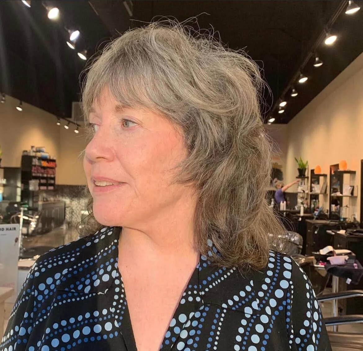 A woman with wavy gray hair and fair skin looking to her left inside a salon with shelves and customers in the background.