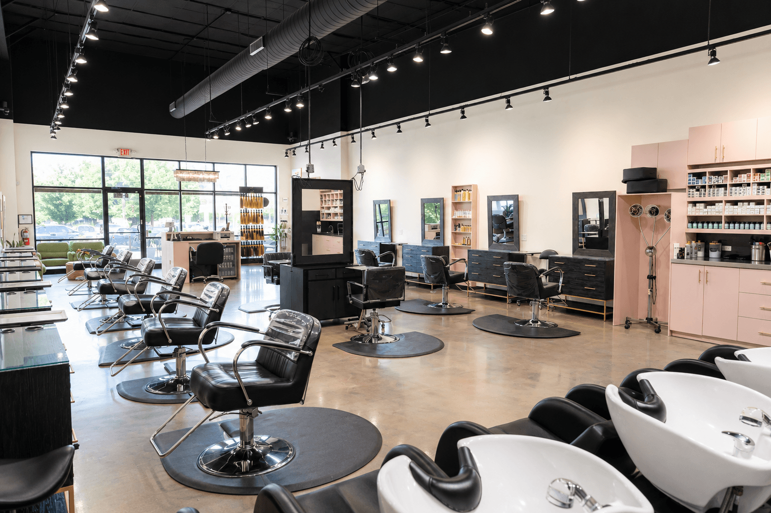 Interior of a modern hair salon with black salon chairs, hair washing stations with white sinks, large windows, and sleek black and pink cabinets.