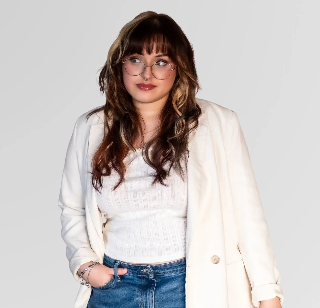 A woman with wavy brown hair, wearing glasses, a white top, and a cream blazer, standing against a plain light gray background.