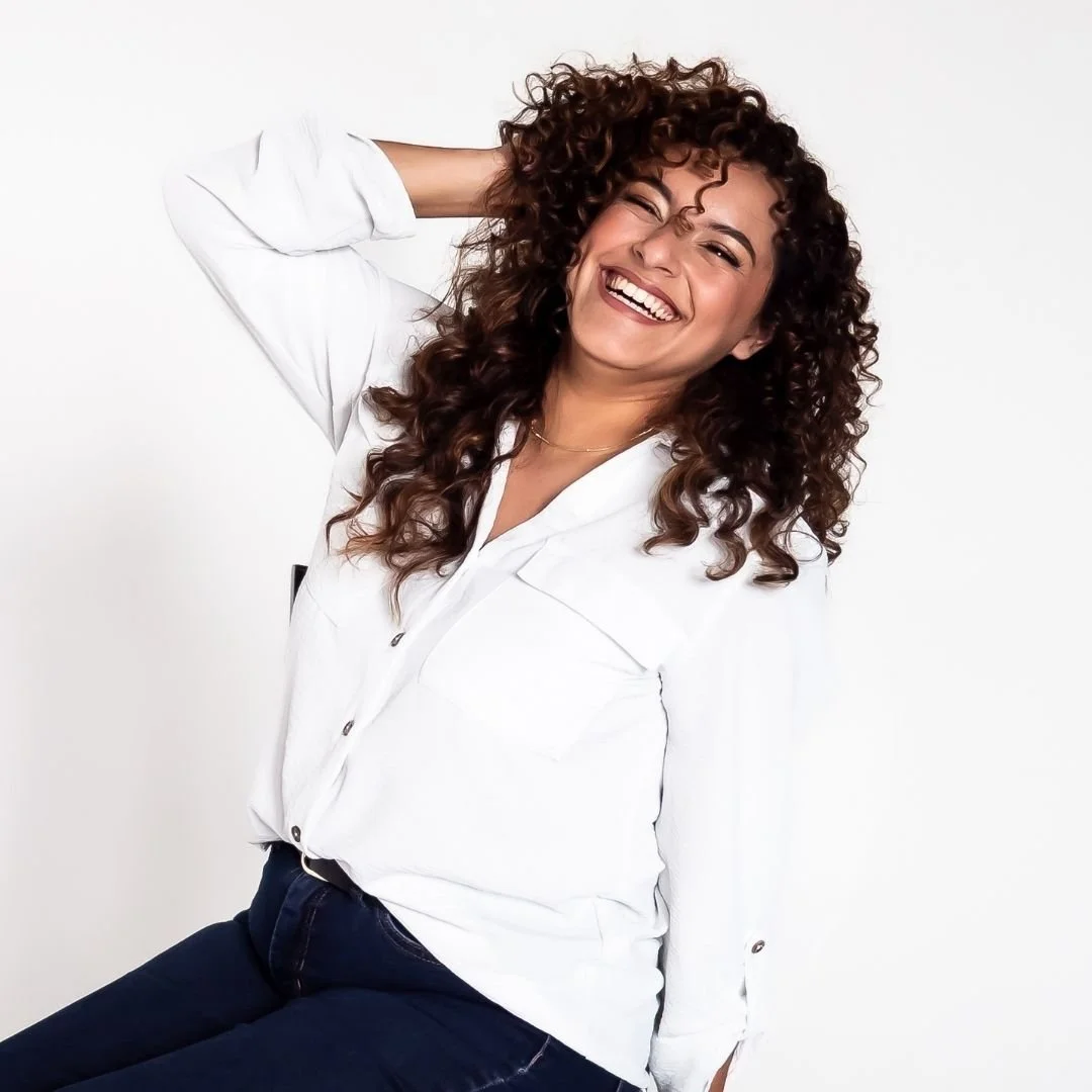 A woman with curly brown hair smiling, wearing a white shirt and dark pants, posing in front of a white background.