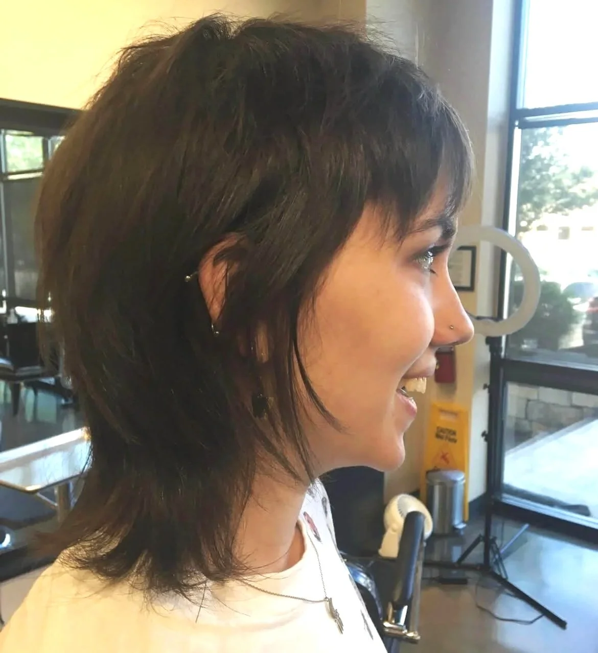 Side profile of a woman with short, dark, layered hair, smiling, inside a salon or similar setting.