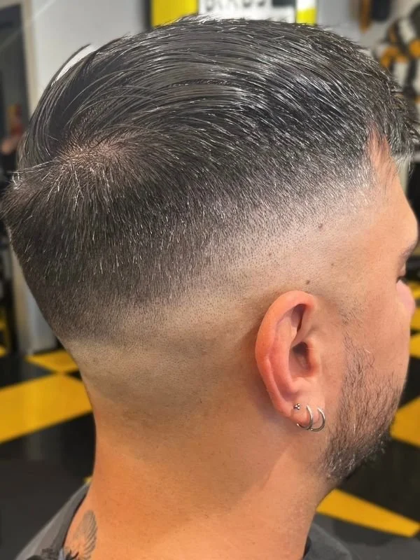 Side view of a man with a fresh skin fade haircut, dark hair on top slicked back, and multiple earrings in his ear.
